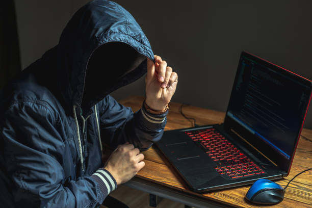 A hacker anonymous in a hood is typing on a laptop keyboard in a dark room. Cybercrime fraud and identity theft stock photo