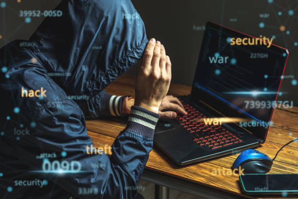 Hacker in a hood with a phone is typing on a laptop keyboard in a dark room. Cybercrime fraud and identity theft stock photo