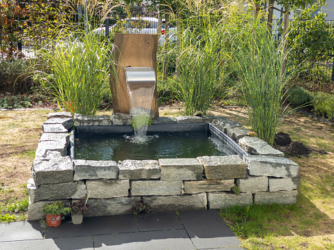 Urban garden with a square high pond built from natural rock with a small waterfall created by a stainless steel outlet which is mounted on a vertical wooden beam, surrounded by green grass