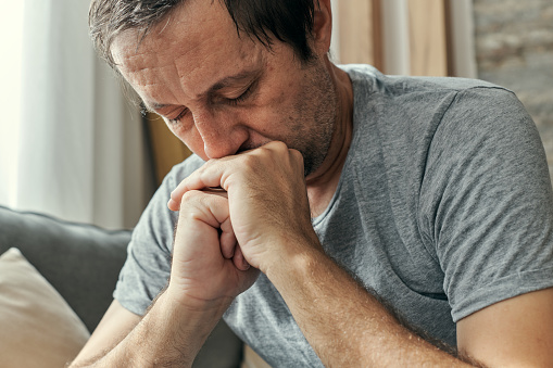 Depressed sulking man sitting alone at living room sofa and thinking, adult caucasian male going through midlife crisis