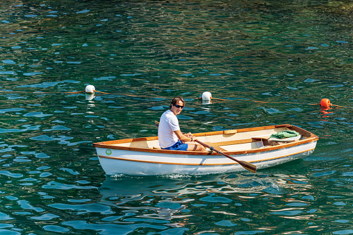 Tellaro, Italy - July 21th, 2019: An adult man (45-49 years) aboard of a small white and brown wooden rowing boat in the blue Mediterranean sea, on a sunny summer day, in front of the ancient Tellaro village, Liguria, Italy, southern Europe.