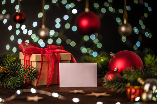 Front view of a gift box beside a blank greeting card with copy space surrounded by christmas balls, pine twigs and christmas lights. Focus on foreground.