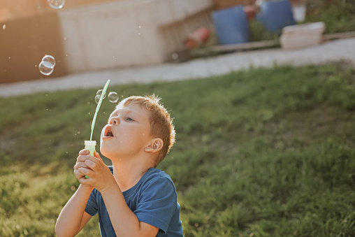 A boy in a blue t-shirt is blowing bubbles in the backyard.