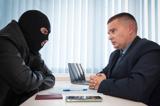 businessman and robbers are sitting at a table. a racketeer in a black balaclava forces to sign a contract. the concept of a raider takeover of the company. signing of important documents. - 偷拍的照片 個照片及圖片檔