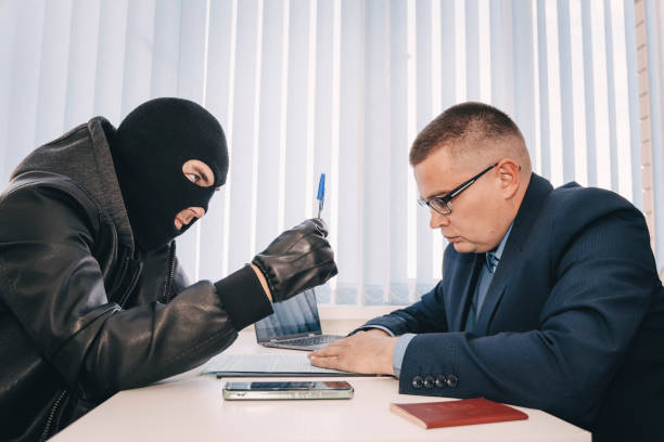 businessman and robbers are sitting at a table. a racketeer in a black balaclava forces to sign a contract. the concept of a raider takeover of the company. dangerous deal - 偷拍的照片 個照片及圖片檔