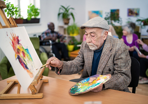 A grey haired retired gentleman is painting a colorful picture on a small easel on a table, with more residents are chatting in comfortable chairs behind him