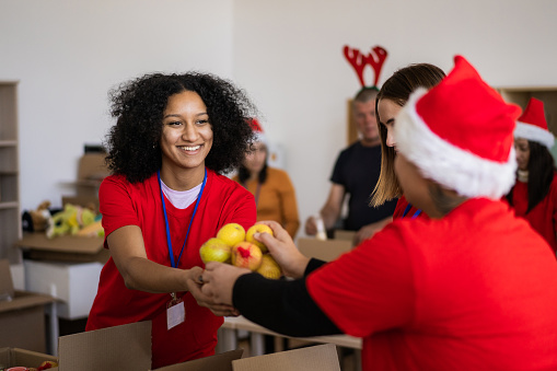 A smiling woman is taking some fresh fruit from another volunteer while they pack and sort donated groceries for distribution during the festive season for impoverished families