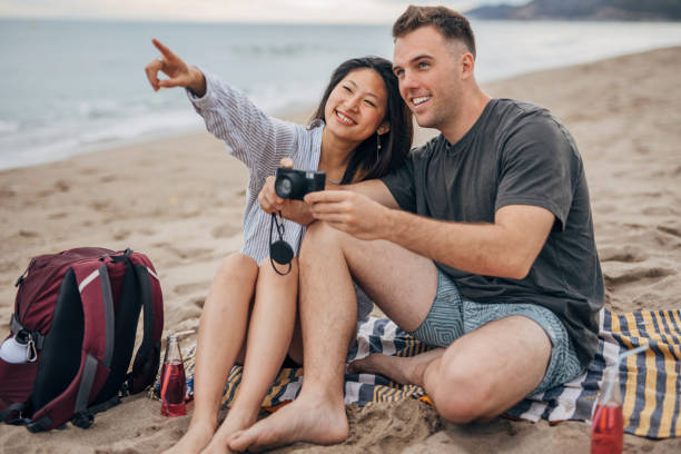 istock Young multi ethnic couple sitting on a beach taking photos 1425148281