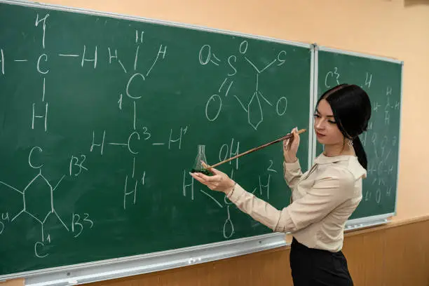 Photo of female student is practicing in a chemistry classroom with flasks on of a board with formulas.