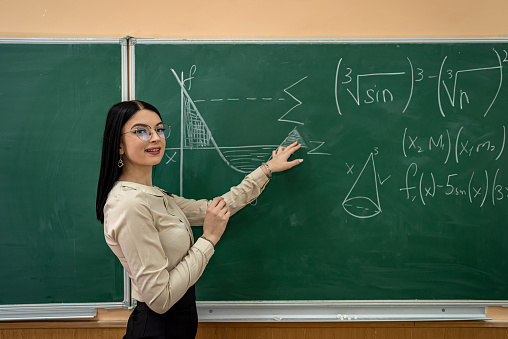 young pretty female teacher writing on a chalkboard completing mathematical equations in the classroom. education concept