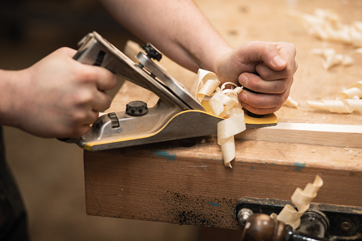 Close-up of cropped carpenter hands working with plane equipment tool on the workbench. Carpentry planer in workshop, garage. Sawdust and shaving chips around on the table. Woodworking industry
