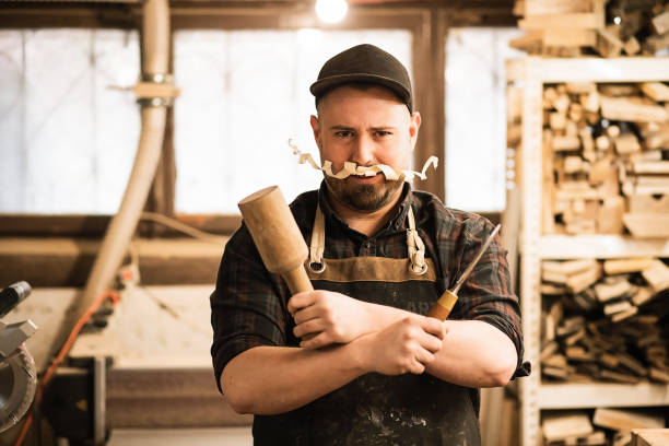 sonriente y divertido trabajo de carpintero masculino en gorra y overol con bigote de afeitar, sosteniendo cincel cruzado y martillo de madera - work tool chisel wood mallet fotografías e imágenes de stock