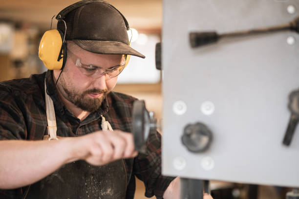 Closeup serious and concentrated man worker, carpenter in protective dirty apron, coveralls, glasses, headphones working with grinder, cutting wood machine with circular saw. Workshop, small business