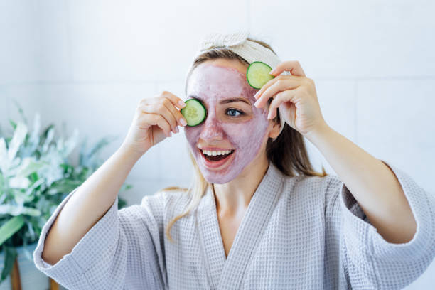 a young smiling woman with pink clay facial mask holds cucumber slices making a refreshing eye mask in bathroom. natural cosmetic procedures for skin care at home. beauty self-care. selective focus. - facial mask spa treatment cucumber human face imagens e fotografias de stock