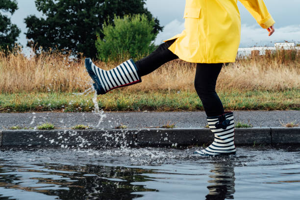 Woman having fun on the street after the rain. Cropped woman wearing rain rubber boots and yellow raincoat walking into puddle with water splash and drops. Fall weather. Selective focus Woman having fun on the street after the rain. Cropped woman wearing rain rubber boots and yellow raincoat walking into puddle with water splash and drops. Fall weather. Selective focus. rubber boot stock pictures, royalty-free photos & images
