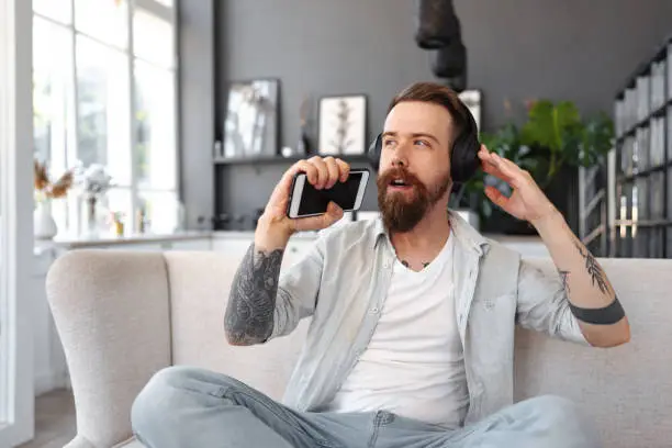 Photo of Positive bearded man listening music with headphones while sitting on couch at home