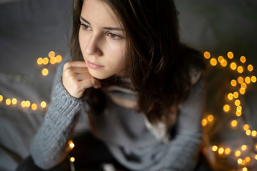 Wearing a warm sweater a young woman is resting her  head on her hand while sitting on a bed surrounded by illuminated Christmas lights
