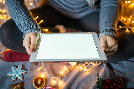 A young woman wearing winter sweater is sitting cross-legged on a bed holding a blank screened digital tablet in front of her, on the bed beside her are illuminated string lights and Christmas decorations