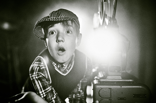 Cute boy old fashioned cinema film projectionist. The boy is standing next to the 16 mm film projector. He is watching cinema with his head on the hand. Studio shooting, black and white image