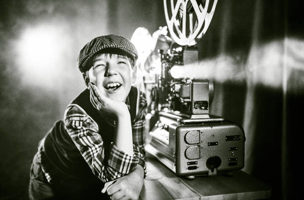 Joyful Boy Film Projectionist In Old-fashion Cap Is Watching Film Using Old Cinema Projector Cute boy old fashioned cinema film projectionist. The boy is standing next to the 16 mm film projector and laughing. He is watching cinema with his head on the hand. Studio shooting, black and white image vintage movie projector stock pictures, royalty-free photos & images