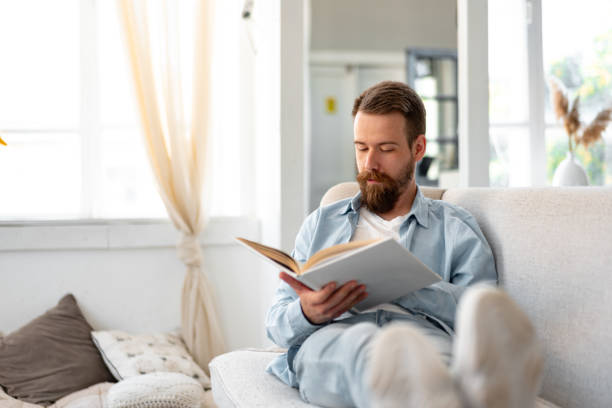 istock Young bearded handsome man reading book on couch at home 1425158164