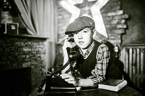 Elementary age boy dressed in vintage clothes is sitting at the table. The boy is talking on the old-fashioned phone. Studio shooting, black and white image, retro style