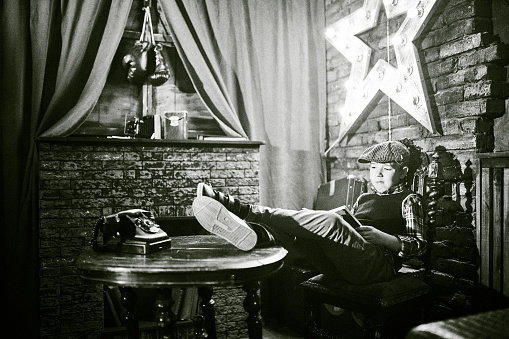 Elementary age boy dressed in vintage clothes is sitting on a chair with his legs on the table. The boy is reading a book. Studio shooting, black and white image, retro style