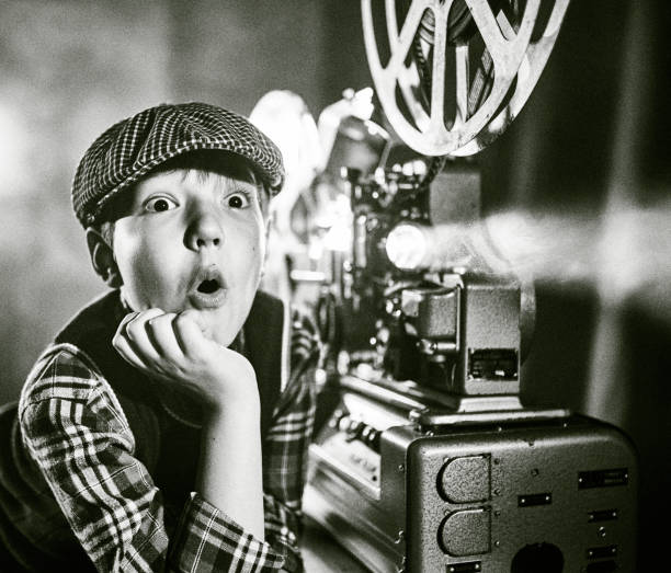 Cute boy old fashioned cinema film projectionist. The boy is standing next to the 16 mm film projector. He is watching cinema with his head on the hand. Studio shooting, black and white image