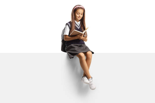 Girl in a school uniform sitting on a blank panel and reading a book stock photo