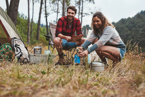 Happy young couple camping outdoors in nature. Cheerful young woman igniting a camp stove with her boyfriend sitting by.