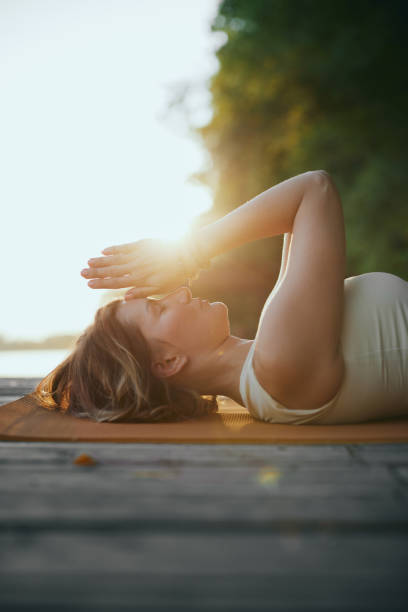 istock A mindful woman is lying on her back and holding hands in a namaste position while practicing breathing on the dock near the river in nature. 1425167978
