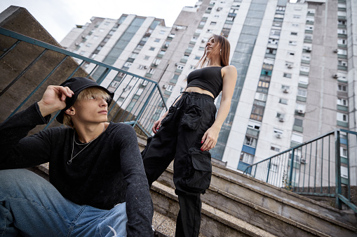 Low angle view of a teenage couple poses in the urban exterior on the stairs surrounded by skyscrapers and buildings.