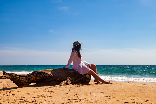 Rear view adult woman in white beachwear, hat and sunglasses, looking away on azure sea, sitting on tropical sandy beach at waves ocean background. Travel tourism vacation concept. Copy text space