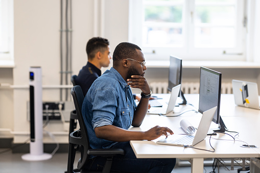 Side view of two multiracial men working on desktop computers at a coworking office. Male professionals sitting at desks and working on computers in open plan office.