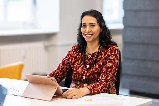 Portrait of a confident mature woman sitting at desk with digital tablet. Middle eastern businesswoman at coworking office looking at camera and smiling.