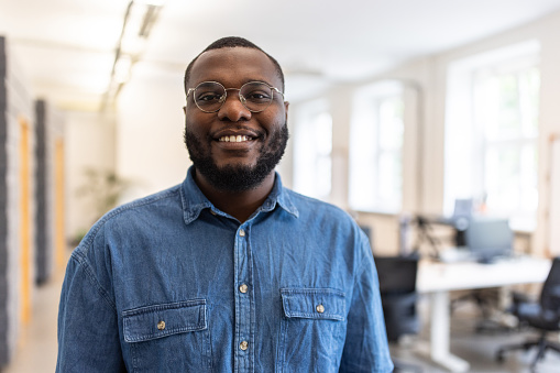 Portrait of young African man in coworking office space looking at camera and smiling. Smiling man with beard and eyeglasses in casuals standing in startup office.