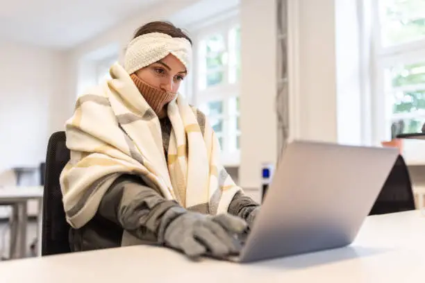 Young woman covered with a blanket using laptop while sitting in office. Sick woman feeling cold wrapped in blanket working at startup office.