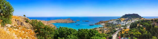 Panoramic view of colorful harbor in Lindos village and Acropolis, Rhodes. Aerial view of beautiful landscape, ancient ruins, sea with sailboats and coastline of island of Rhodes in Aegean Sea