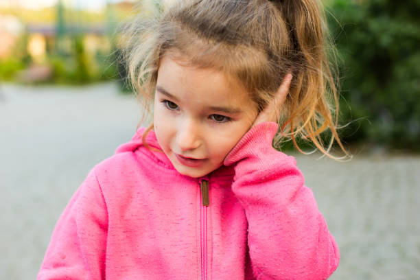 ein kleines mädchen in einem rosa kapuzenpullover mit einem traurigen und tränenreichen gesicht hält ihr ohr. ohrenschmerzen, mittelohrentzündung, schwellung der wange, zahnfleisch, zahnschmerzen, kinderchirurgie, hno-heilkunde. kindermedizin - chewing gum audio stock-fotos und bilder