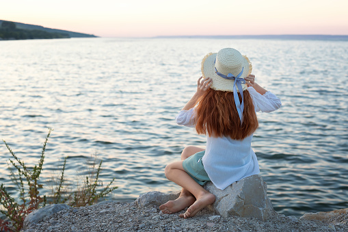 A beautiful red-haired girl in a hat sits on a stone on the banks of a river, lake, sea. The girl is alone on the street. The girl is sitting on the rocks. Lonely man. Rest on the sea. The girl is thinking on the beach. Back to the camera.