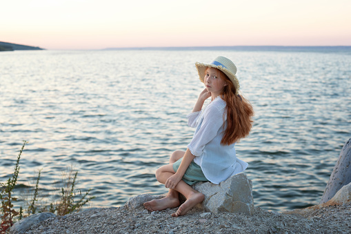 A beautiful red-haired girl in a hat sits on a stone on the banks of a river, lake, sea. The girl is alone on the street. The girl is sitting on the rocks. Lonely man. Rest on the sea. The girl is thinking on the beach. Looks into the camera.