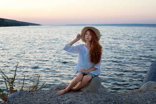 A beautiful red-haired girl in a hat sits on a stone on the banks of a river, lake, sea. The girl is alone on the street. The girl is sitting on the rocks. Lonely man. Rest on the sea. The girl is thinking on the beach.