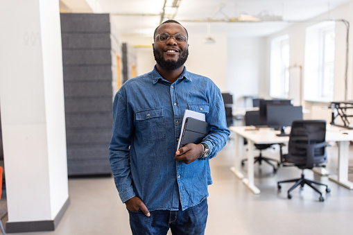 Portrait of confident young African man at office looking at camera and smiling. Smiling man with beard and eyeglasses standing in startup office.