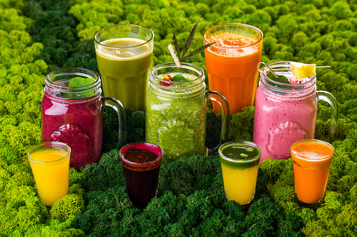 Three jars with handles and fruit smoothies made from strawberries, raspberries and kiwi, fruit wedges and hemp litter. Stand against a background of stabilized moss. Four glasses with carrot, beet, celery and sea buckthorn juice on a background of stabilized moss. Glass with fresh carrot, glass with fresh apple.