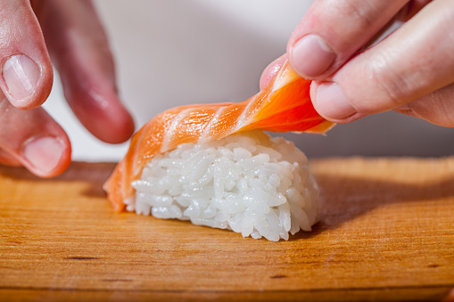 Male hands are laying salmon fillets on sushi, sushi is on a wooden board.
