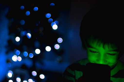 Child being illuminated by the led light produced by the smartphone and in the background, and out of focus, lights coming from a Christmas tree. Technological Christmas concept.