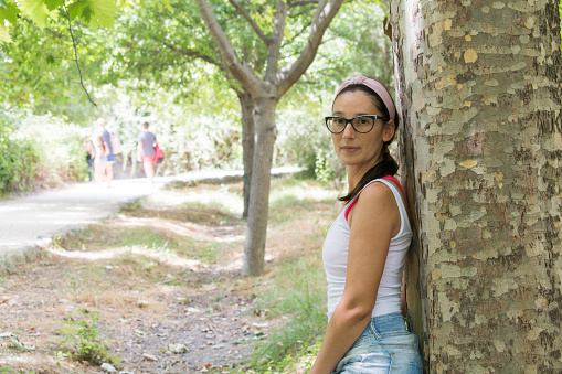 A young freckled woman with glasses leans against a tree with a family out of focus behind, Fuentes del Marques, Caravaca de la Cruz, Murcia, Spain