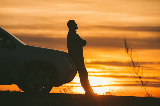 A silhouette of a man beside his vehicle having mechanical difficulty. SUV. Roadside assistance needed. Car trouble. Horizontal colour image. Rural highway. Themes include mechanical breakdown, cars, automotive, repair, towing, trouble, highway, roadside, hood, engine trouble, vehicular breakdown, mechanical, suv, driving, motoring, auto, auto insurance, auto, mechanic, and alone. 