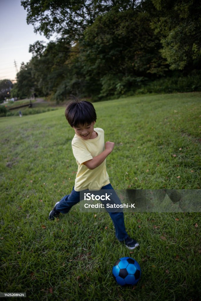 Little boy kicking a ball in public park Japanese family visiting public park by bicycle to enjoy walking in forest, playing with ball and equipments, eating foods. 6-7 Years Stock Photo