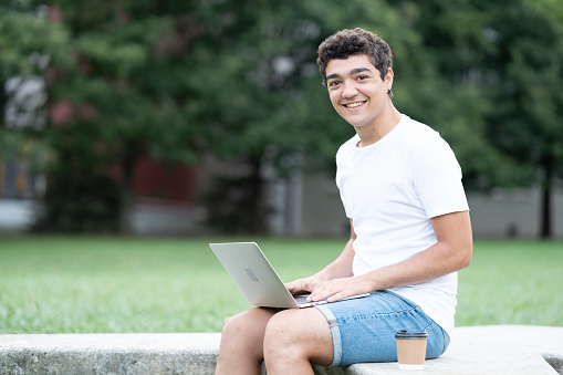 Hispanic freelancer teen boy working on laptop, looking and smiling at camera outdoors in a park.